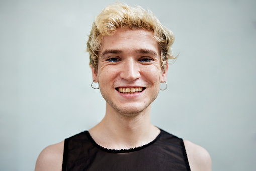 Head and shoulder view of shorthaired member of LGBTQIA community wearing black tank top, earrings, and smiling at camera.