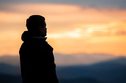 A captivating scene as a teenage boy, wearing a black rain hoodie, stands as a striking silhouette against the backdrop of a breathtaking and colorful sunset sky in the mountainous wilderness. The warm hues of the setting sun create a serene and awe-inspiring moment, capturing the beauty of nature's transition from day to night.