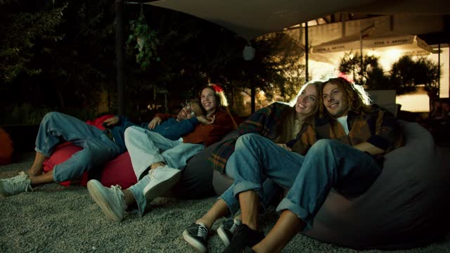 Two couples in love are sitting on beanbag chairs and watching a movie. A party outside the city. Rest in the country house