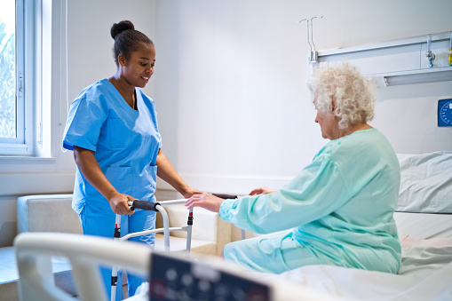 Smiling female nurse talking with patient sitting on bed in hospital ward, holding mobility walker.