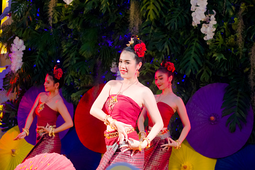 Capture of happy young thai women Rum Thai dancer standing in elegant pose. Captured  in Chiang Mai. Women are dressed  elegant in traditional clothes in front of umbrella decor.  Women are red dressed and are dancing classical Rum Thai.  Performance is on a small stage and they are dancing in public for people who are visiting market as part of several entertainment of dancers and singers. Scene is on night market at One Nimman square in Chiang Mai.