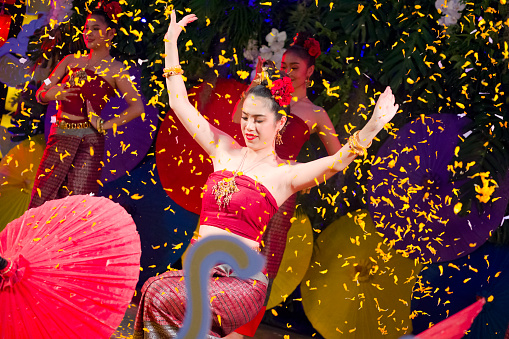 Happy young thai women Rum Thai dancer under flower petal rain. She is  stretching out arms standing in elegant pose at end of dance performance. Dance is captured  in Chiang Mai. Women are dressed  elegant in traditional clothes. In background are different colored paper  umbrellas as  decor.  Women are red dressed and are dancing classical Rum Thai.  Performance is on a small stage and they are dancing in public for people who are visiting market as part of several entertainment of dancers and singers. Scene is on night market at One Nimman square in Chiang Mai.