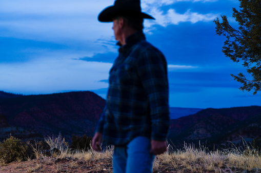 Cowboy Rancher Looking Off into Distance - Side profile shot of blurred OOF rancher cowboy wearing cowboy hat with focus on mid ground captured in a natural outdoor Western USA area with mountain backdrop. Rancher keeping an eye on his cattle, captured at dusk during blue hour.