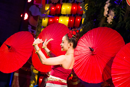 Portrait of young thai woman  dancing classic  Rum Thai dance in public. Live performance at night done by  women who are dancing in a group with red paper umbrellas and red and yellow lights decor in background. Women are red dressed and are dancing classical Rum Thai.  Performance is on a small stage and they are dancing in public for people who are visiting market as part of several entertainment of dancers and singers. Scene is on night market at One Nimman square in Chiang Mai. Women are dressed  elegant in traditional clothes.