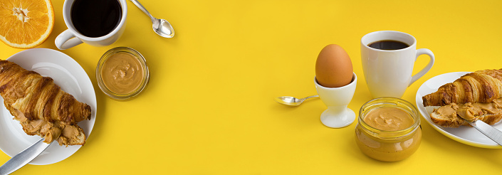 Breakfast with croissant, peanut paste, black coffee and boiled egg on the yellow background. Copy space. Close-up.