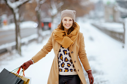 smiling modern female in brown hat and scarf in camel coat with gloves and shopping bags outside in the city in winter.