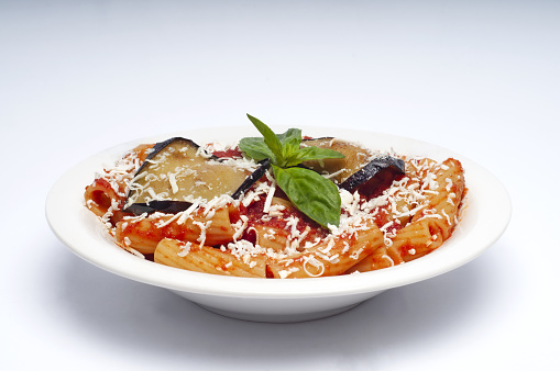 Pasta Alla Norma. Delicious Sicilian pasta dish with roasted eggplant, tomato sauce, grated ricotta and fresh basil served in a white ceramic bowl. White background, selective focus, horizontal.