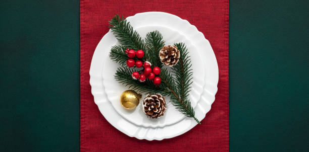 Christmas table with decorations, white empty plate on red tablecloth, green background, top view, above. Celebration xmas eve: art arrangement, red accents. Fir cones, evergreen tree. Holiday banquet