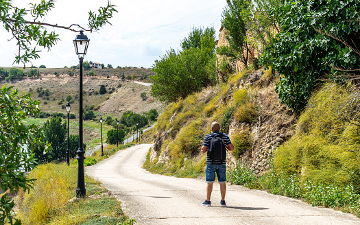 Middle-aged tourist with a backpack goes sightseeing in a rural area with a hill - backpacker walks on an asphalt road where there is a steep slope, looks at the landscape in Maderuela in the province of Segovia - Spain.