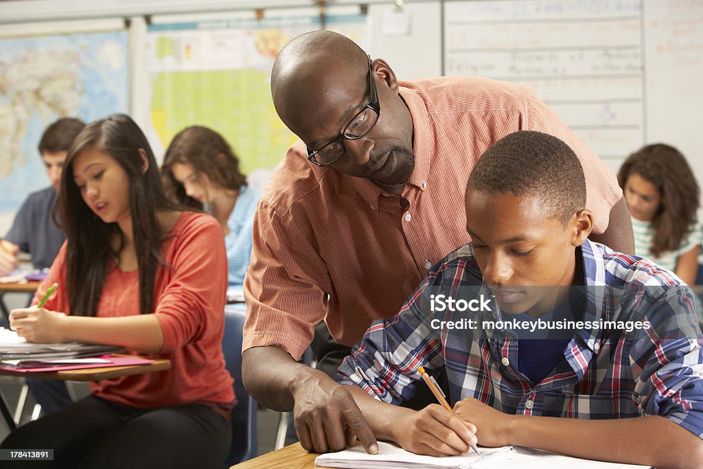 Teacher helping a male pupil studying at a desk in classroom Teacher Helping Male Pupil Studying At Desk In Classroom Looking Over Shoulder Teacher Stock Photo