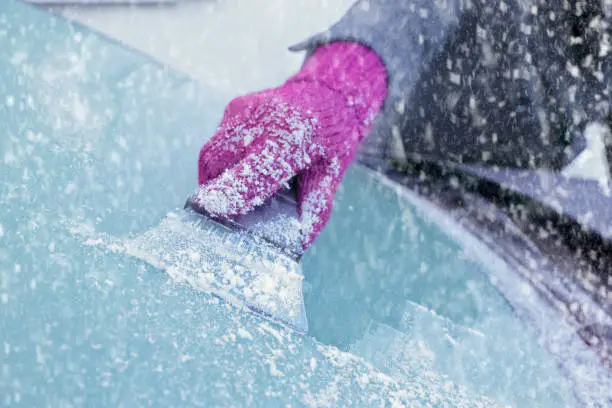Close-up of a woman's gloved hand using an ice scraper to remove ice from the frozen windscreen of her car before driving