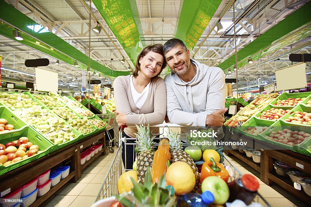 Happy customers Image of happy couple with cart full of products looking at camera in supermarket Supermarket Stock Photo