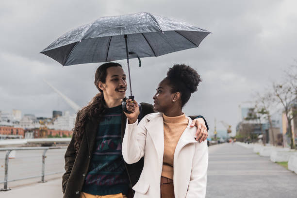 giovane coppia che parla e cammina usando l'ombrello a puerto madero, buenos aires - candid women african descent umbrella foto e immagini stock