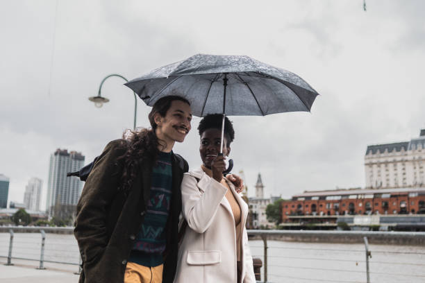 giovane coppia che parla e cammina usando l'ombrello a puerto madero, buenos aires - candid women african descent umbrella foto e immagini stock