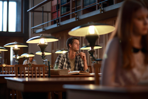 Young male reading an old book with an old electric lamp in a library's reading room at night.