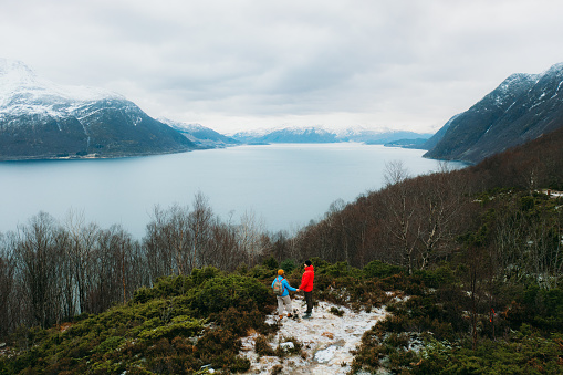 Drone panoramic photo of heterosexual couple wearing blue and read jacket hiking with their dog to the top of the mountain peak looking at the dramatic ocean and snowcapped mountain peaks during scenic sunset in the Western Norway, Scandinavia