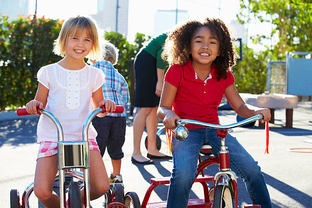 Two Girls Riding Tricycles In Playground Smiling At Camera Happy