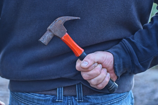 a male criminal in gray clothes holds a large brown hammer in his hand behind his back on the street