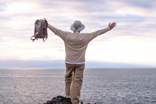 Back view of adult senior man with hat and open arms in front to the sea holding backpack looking at horizon over water. Freedom and travel concept. Copy space