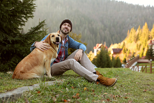 Happy man and adorable dog sitting on green grass in mountains. Traveling with pet