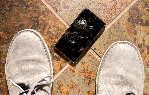 A smartphone lies broken between the shoes of its owner just after being dropped.