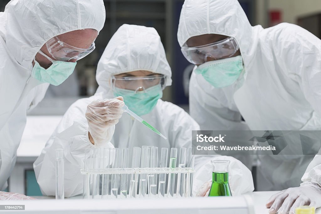 Chemist adding green liquid to test tubes Chemist adding green liquid to test tubes with two other chemists watching in the lab Medical Clinic Stock Photo