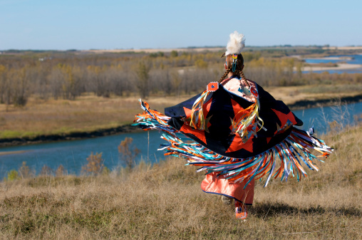 A woman performing a First Nations fancy shawl dance in a field alongside the river in Saskatoon, Saskatchewan