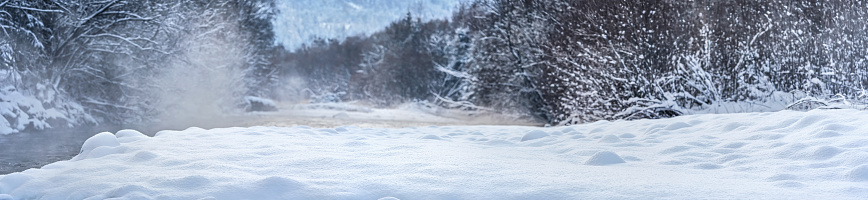 Cold winter river, steam visible above water, dark trees on both sides, fresh snow covered shore in foreground - wide panorama