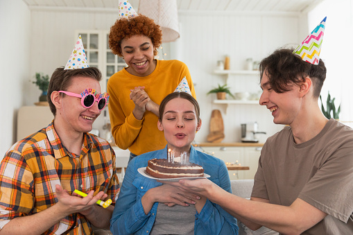 Make a wish. Woman wearing party cap blowing out burning candles on birthday cake. Happy Birthday party. Group of friends wishes girl happy birthday. People celebrating birthday with party at home