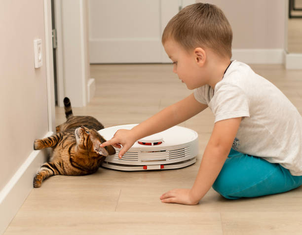 a little boy and a beautiful domestic leopard cat of the bengal breed have fun playing in a home interior with a robot vacuum cleaner that cleans the apartment. - child domestic cat little boys pets imagens e fotografias de stock