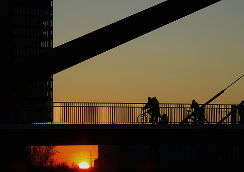 This photo was shot in late July 2023, in Dusseldorf, Germany. Here is depicted a dramatic sunset, contrasting with the silhouettes of of a small group of people going through a pedestrian overpass, and with the silhouettes as well from nearby buildings.