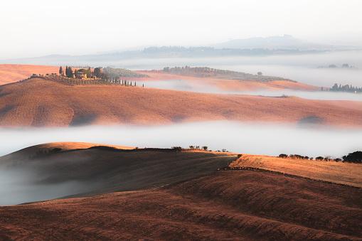 A lone farmhouse on a hilltop peeks through low cloud and fog in the scenic countryside landscape of rolling hills and farmland in rural Tuscany, Italy.
