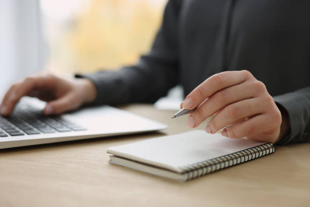 istock Woman with notebook and pen working on laptop at wooden table, closeup. Electronic document management 1784518930
