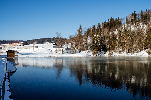Panoramic view of the winter ice-free lake, a bath is built next to the shore, the other part of the coast is covered with larches and pines. Active rest on the lake, holidays in winter, the sun is brightly reflected from the snow surface. High quality photo