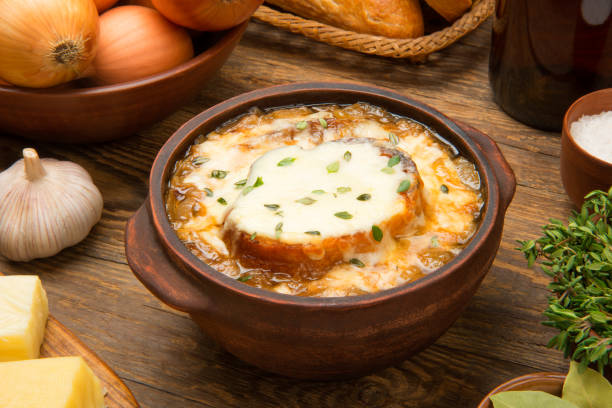 French onion soup in a brown ceramic bowl on a rustic wooden table with ingredients. stock photo