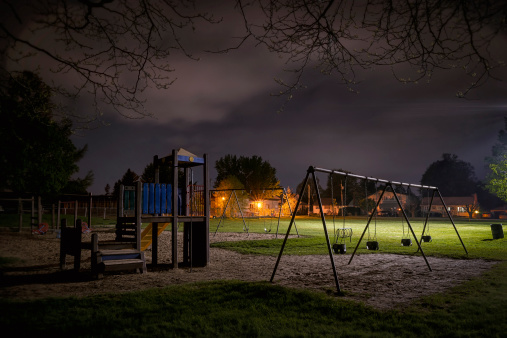 A creepy scene of a deserted children's playground in a suburban park at night time.