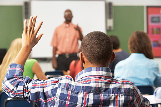 Male Pupil Raising Hand In Class Male Pupil Raising Hand In Class Sitting At Desk hand raised classroom student high school student stock pictures, royalty-free photos & images