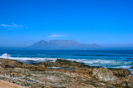 Waves dash against a rocky beach in a seascape at Milnerton Beach in Cape Town, South Africa. Across Table Bay, in the background, is Table Mountain, a major landmark in the city: one can easily navigate around Cape Town, just by looking towards the mountain. Photo shot in the afternoon sunlight.  Horizontal format; copy space.