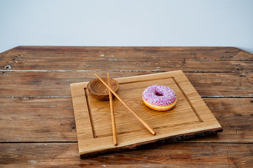 Kitchen equipment made of high-quality material. A bright pink donut, a wooden bowl for sauce with Chinese sticks are located on a rectangular wooden board. High quality photo