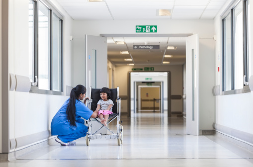 Young female child patient in wheelchair sitting in hospital corridor with Indian Asian female nurse