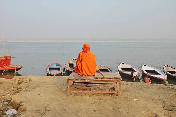 "A holy indian Sadhu with traditional orange clothing is sitting on a wooden bank and looking at the holy river Ganges in Varanasi, Uttar Pradesh, India."