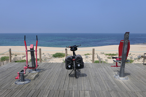 Sports equipment to the left and right of the touring bike in front of a wonderful, blue sea backdrop with a beach in front.
