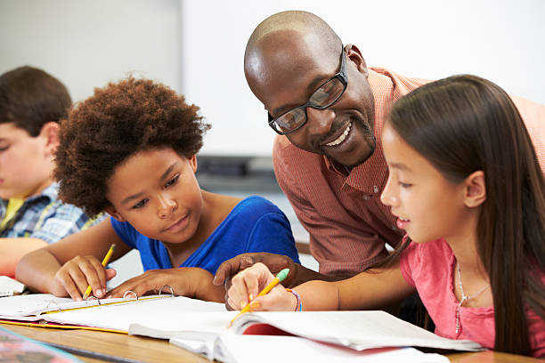 Teacher helping pupils study in the classroom Teacher Helping Pupils Studying At Desks In Classroom Checking Work Smiling elementary age stock pictures, royalty-free photos & images
