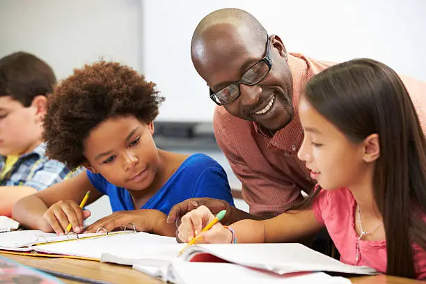 Teacher Helping Pupils Studying At Desks In Classroom Checking Work Smiling