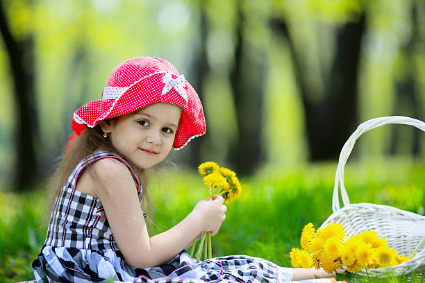 girl with long hair sitting on the green lawn. Beside her is a white basket with yellow dandelions, there is a red hat on her head