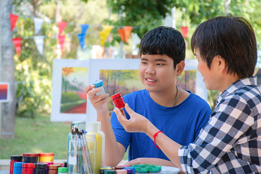 Asian schoolboys and female art teacher is taking outdoor art lesson in poster color drawing, painting and coloring on table in schoolpark, hobby, freetimes activity and learning by doing concept.