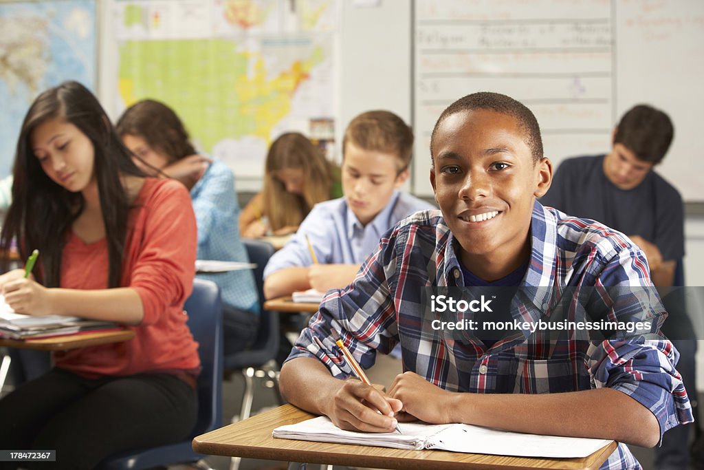 Student writing on notebook in classroom Portrait Of Male Pupil Studying At Desk In Classroom Looking To Camera Smiling High School Student Stock Photo
