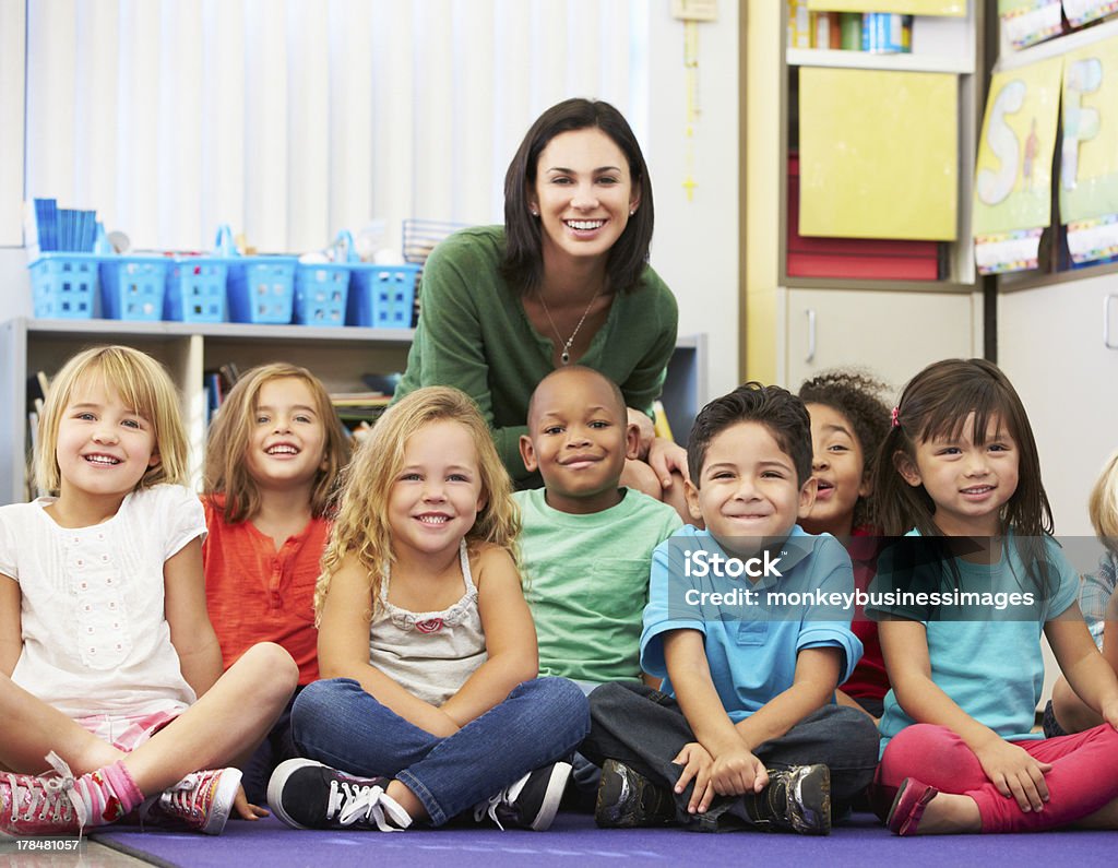 Group of Elementary Pupils In Classroom With Teacher Group of Elementary Pupils In Classroom With Teacher Sitting Down Smiling At Camera Teacher Stock Photo