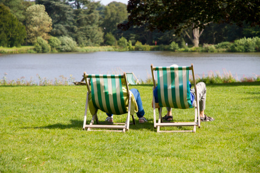 Rear view of elderly couple relaxing on deck chairs in English country park.