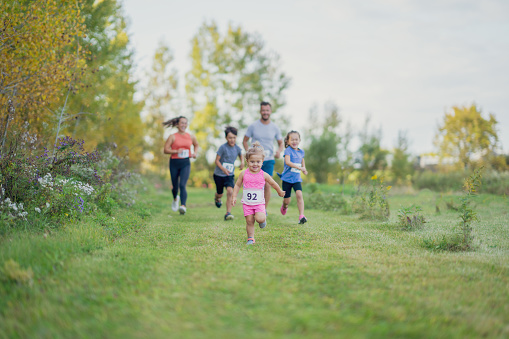 A small young mixed race family is seen participating in a race together as they run outdoors on a warm fall day.  They are each dressed comfortably in shorts and t-shirts and have race bibs on.